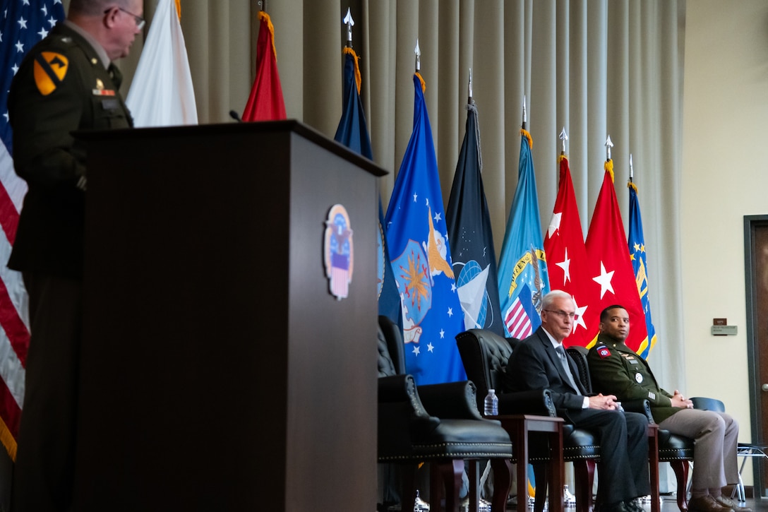 A man wearing a military uniform stands on stage behind a lectern speaking to two other men seated in chairs on the stage.