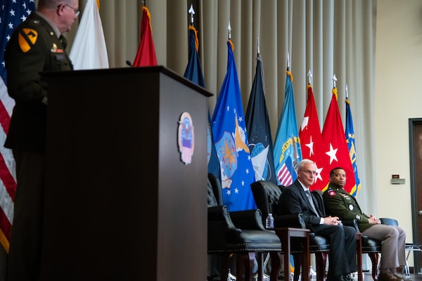A man wearing a military uniform stands on stage behind a lectern speaking to two other men seated in chairs on the stage.