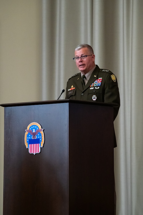 A man wearing a military uniform stands on stage behind a lectern speaking.