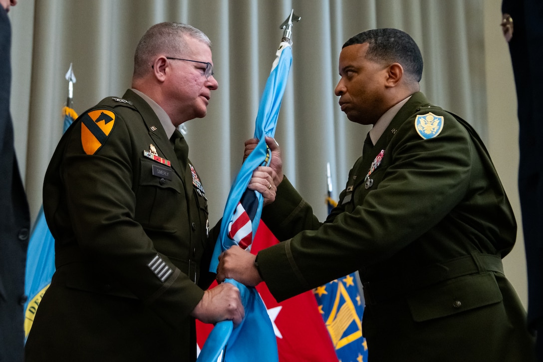 Two men wearing military uniforms hold a blue flag on a flagpole.