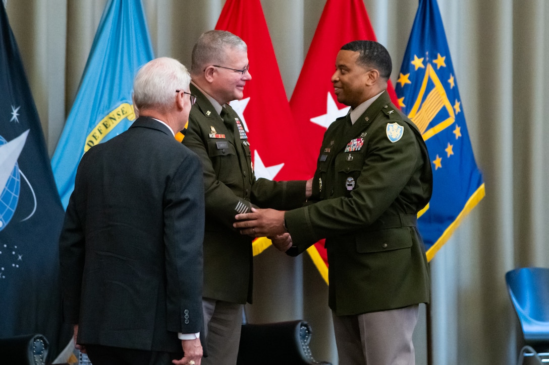 Two men wearing military uniforms stand on a stage shaking hands with a man wearing a suit standing to the side looking at them.