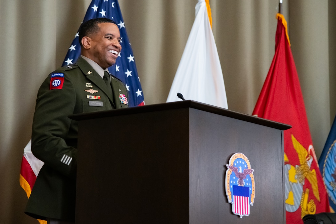 A man wearing a military uniform stands on stage behind a lectern smiling.