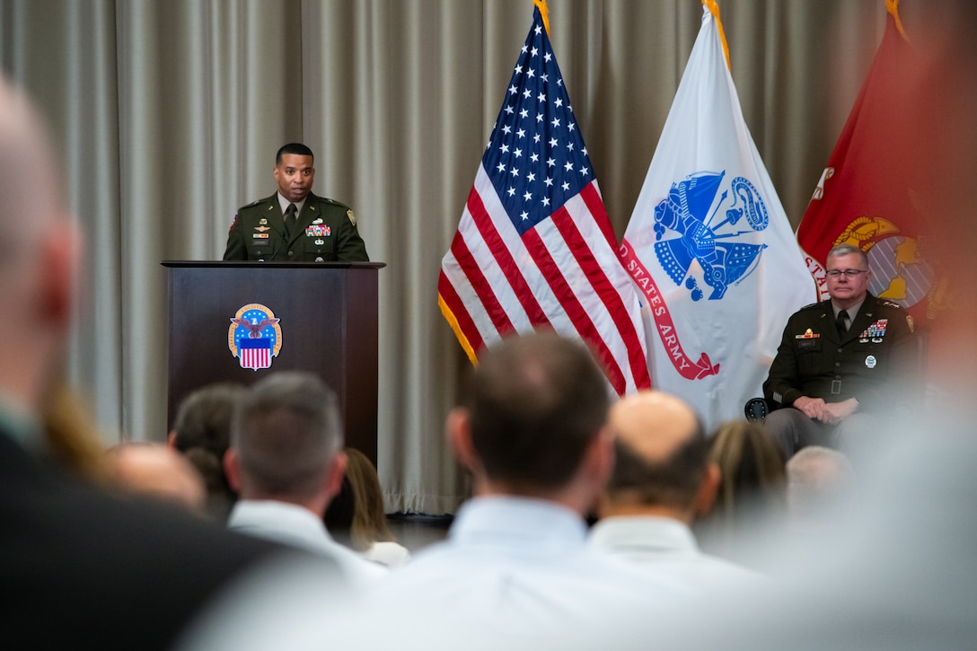 A man wearing a military uniform stands on stage behind a lectern speaking to a crowd.