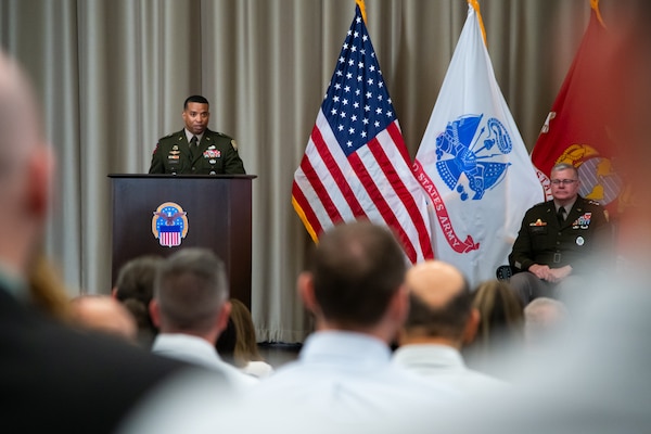 A man wearing a military uniform stands on stage behind a lectern speaking to a crowd.