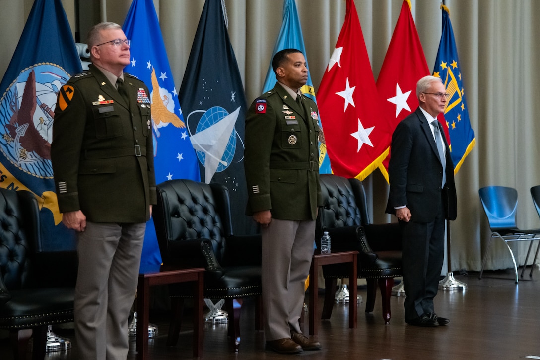 Three men stand on a stage in front of military service flags and two of them are wearing military uniforms.