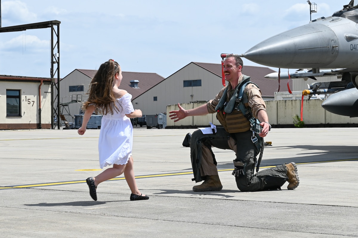 A child runs to a fighter pilot kneeling on the ground with open arms.