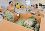 Two women in military uniforms stand in a warehouse in front of a large cardboard box, inside the box are military duffle bags.