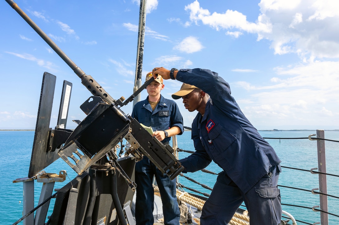 A sailor holds and inspects a large machine gun attached to the deck of a ship as another sailor looks on, with a blue sky and water in the background.