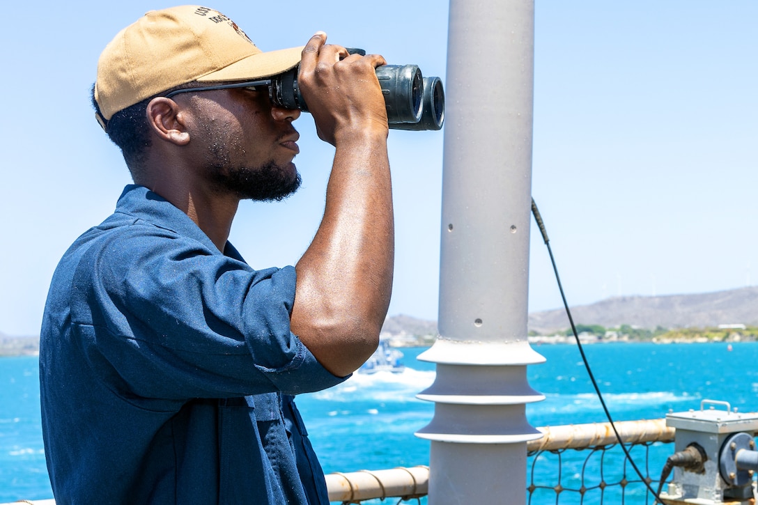 A sailor wearing a blue shirt and hat uses binoculars to look out from a ship, with blue water in the background and hills in the distance.