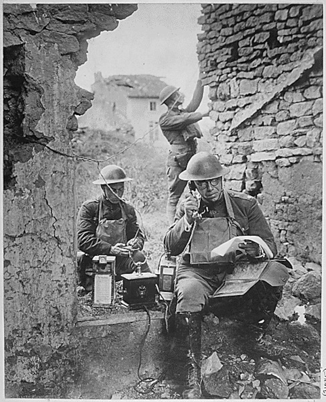 A person sitting in a ruined building puts a rotary telephone to their ear while two people work behind.