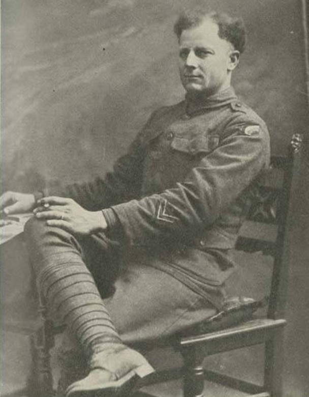 A man in uniform sits in a chair and smokes a cigarette while his right leg is crossed over his left knee.