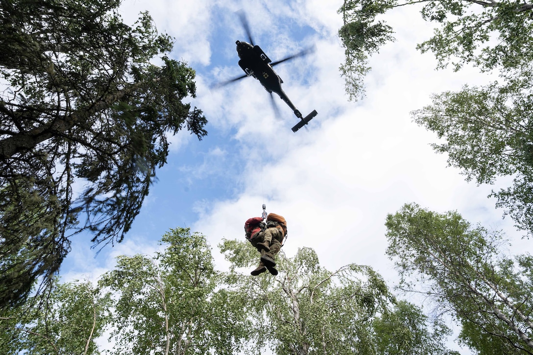 Two people attached to a cable are hoisted toward a helicopter hovering above a wooded area.