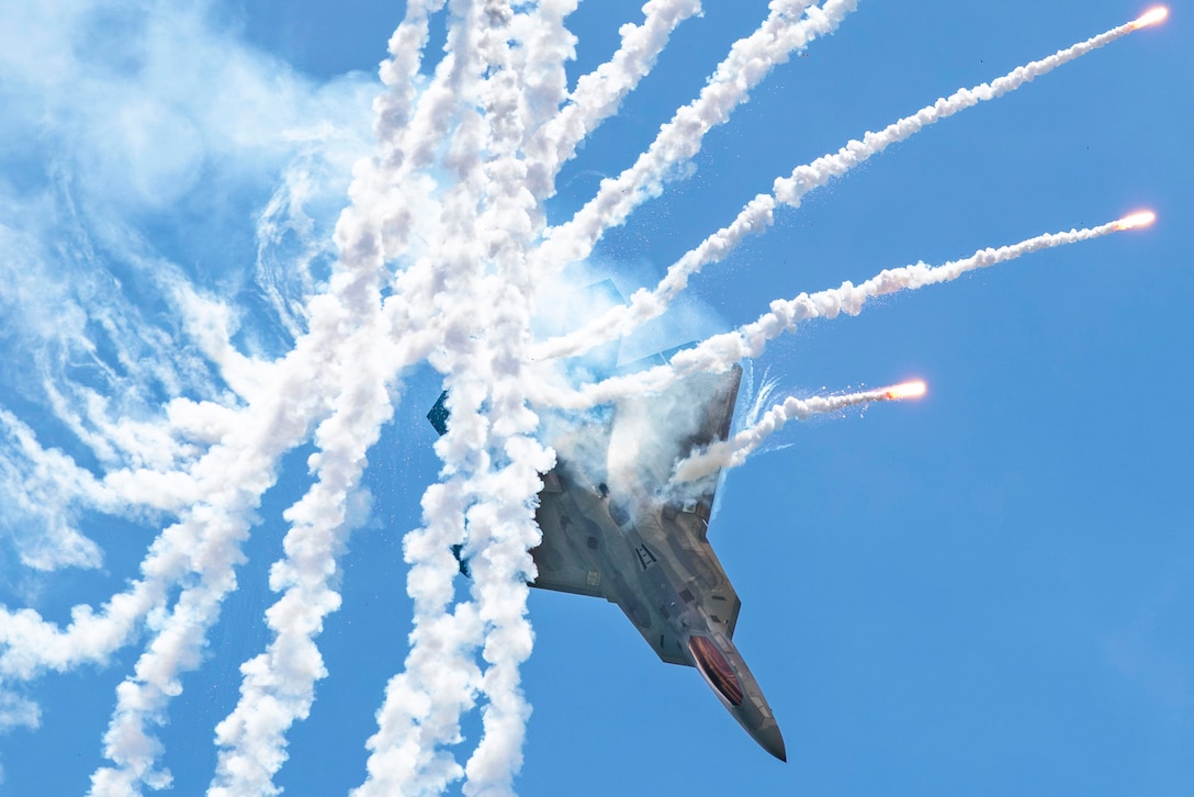 An airman pilots a fighter jet seen from below in a cloudless sky and releases flares, creating smoky trails.