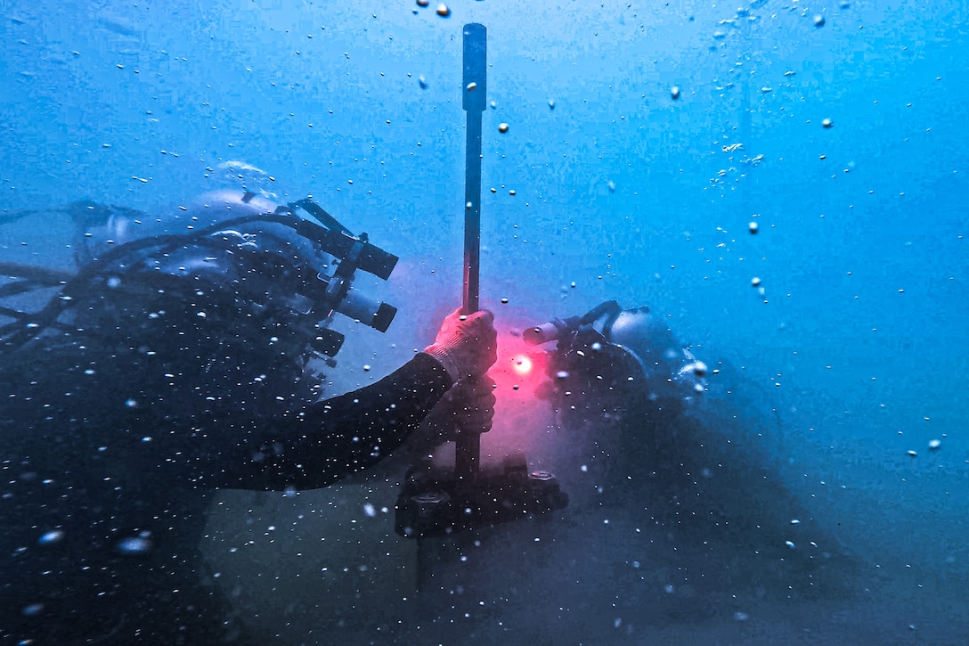 Two Navy divers under blue water and in scuba gear pull a rod from the seafloor as bubbles move around them.