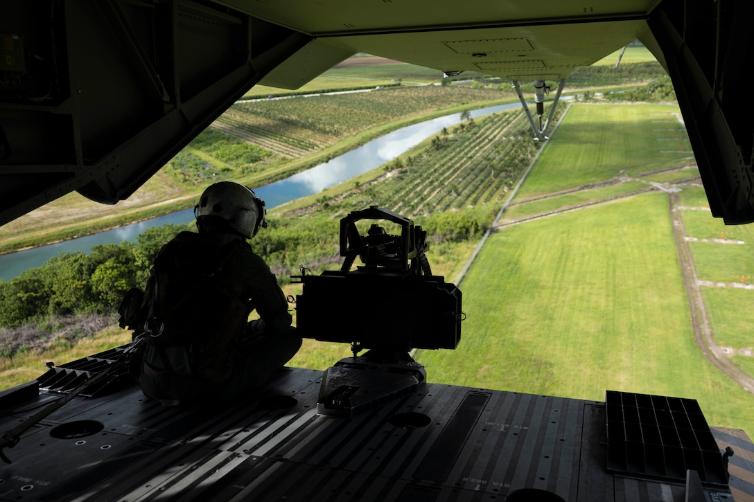 A Marine looks out from the open rear door of a helicopter, with green fields and a river seen below.