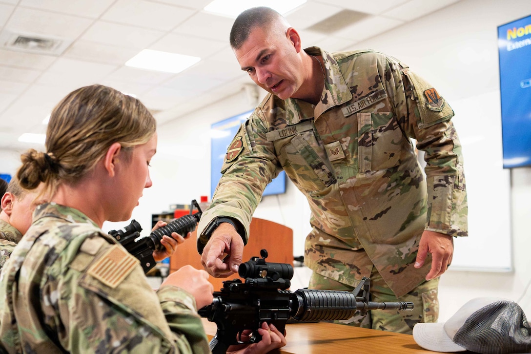 An airman stands and points to a section of a rifle being held by another airman in a classroom setting; other airmen sit nearby.