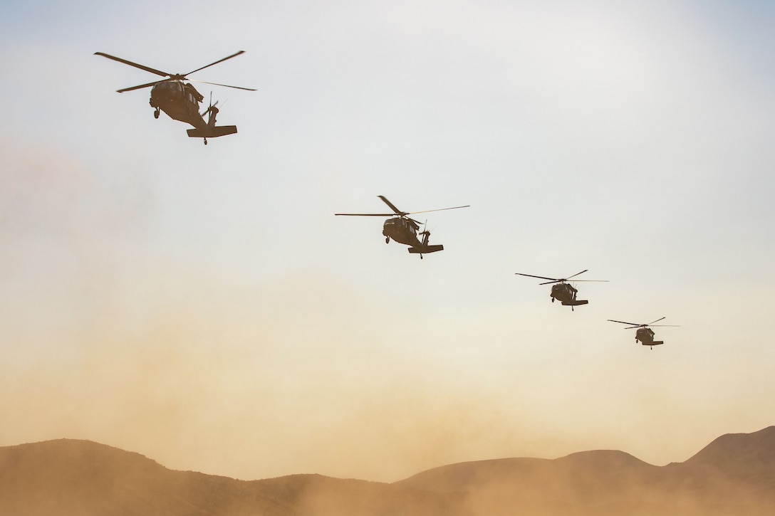 Four helicopters descend toward desert-like terrain, kicking up dust above the land, with hills in the distance and a cloudless sky above.
