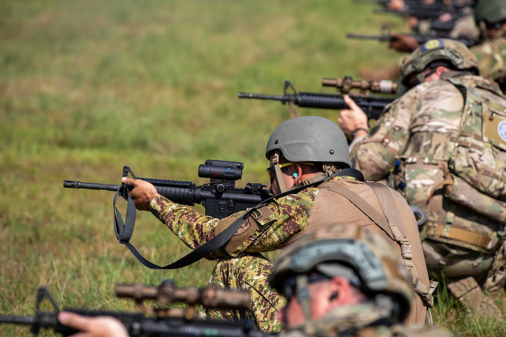 Soldiers squatting in a row and wearing tactical gear aim and fire rifles at a grassy field on a sunny day.