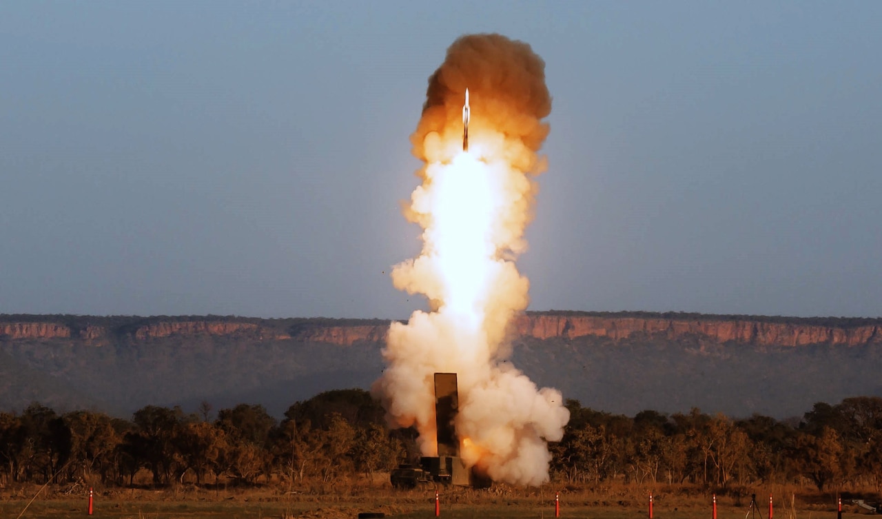 A rocket launches from the ground, leaving a smoke and fire trail in the air. Mountains are in the background.