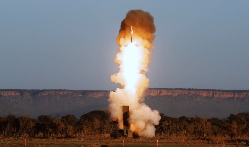 A rocket launches from the ground, leaving a smoke and fire trail in the air. Mountains are in the background.