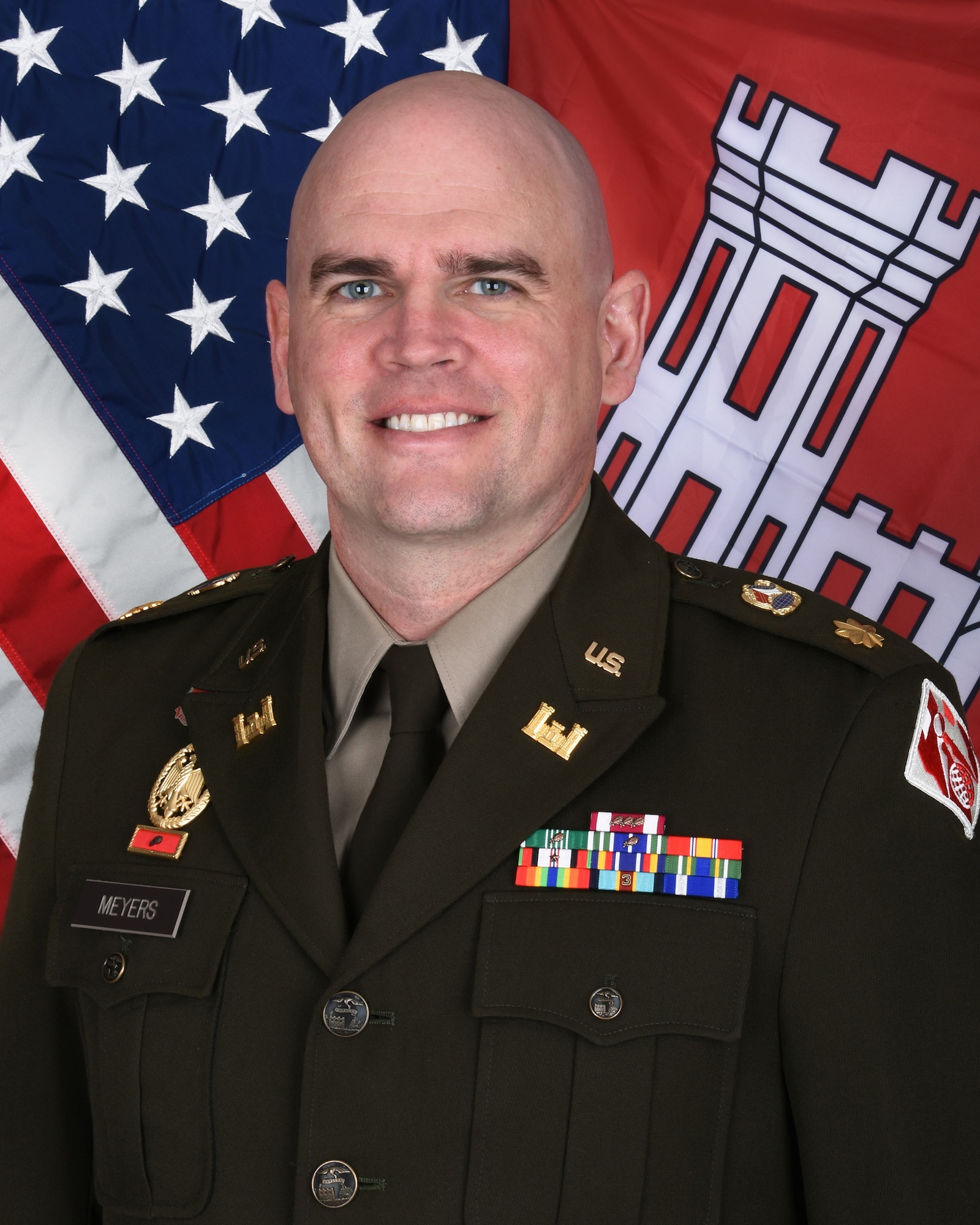 A man in a military dress uniform smiles in front of the American flag.