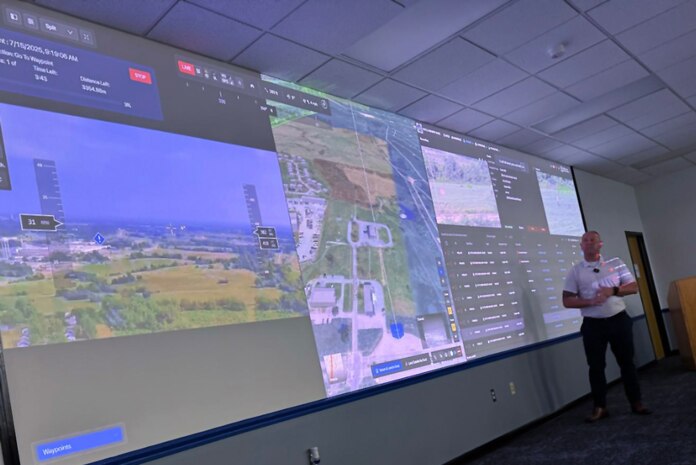 Photo of a man standing in front of a large screen with data and camera views from drones shown in the background.