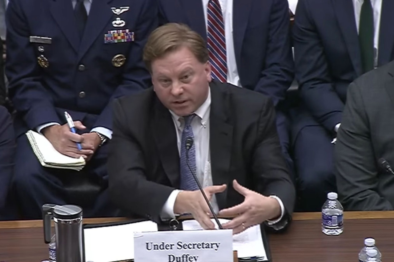 A man in a suit seated at a desk speaks as two people sit beside him and others are seated behind him.