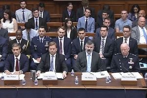 Four men in suits and one in a formal military uniform are seated at a long wooden desk, and others are seated behind them.
