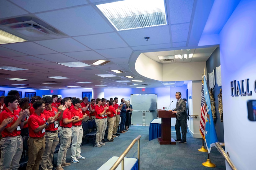 A group of students in red polos stand an clap while Defense Secretary Pete Hegseth watches from a podium.