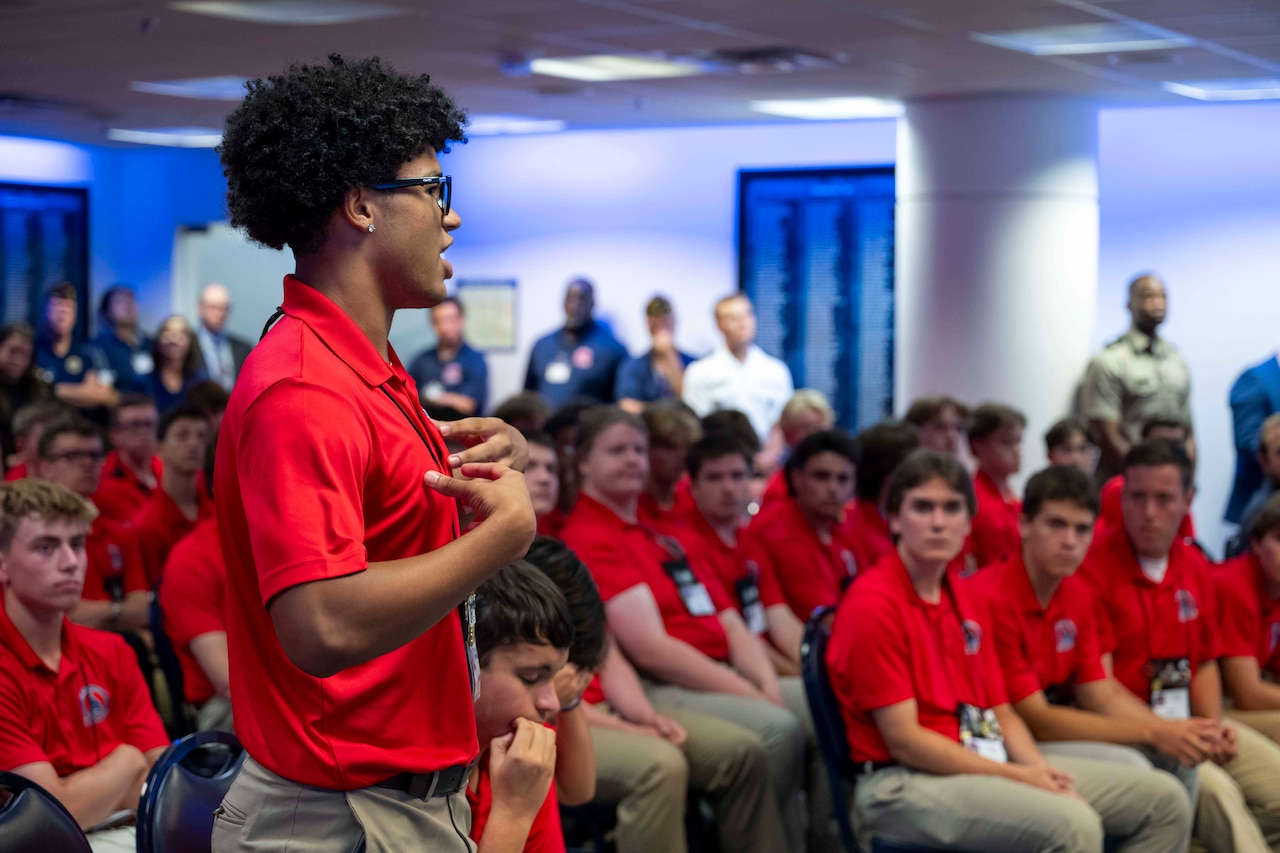 A students in a red polo stands in front of seated students in red polos in a room.