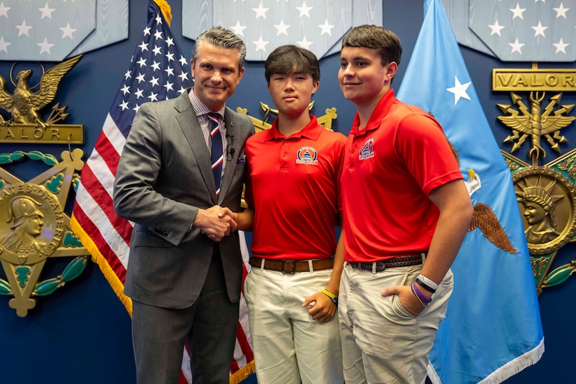 Defense Secretary Pete Hegseth poses with two students in red polos in front of standing flags.