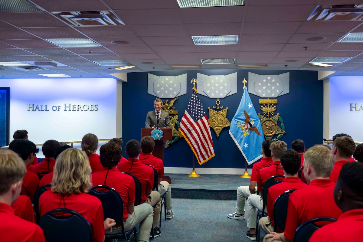 Defense Secretary Pete Hegseth stands at a podium and speaks to a group of seated students in red polos inside the Hall of Heroes.