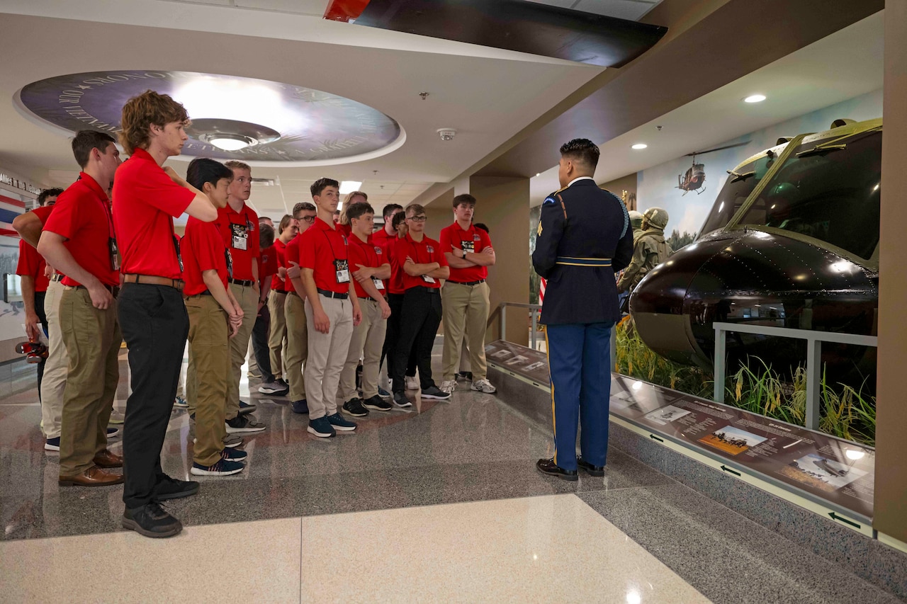 A group of students in red polos listen to a service member standing in front of a helicopter display in the Pentagon.