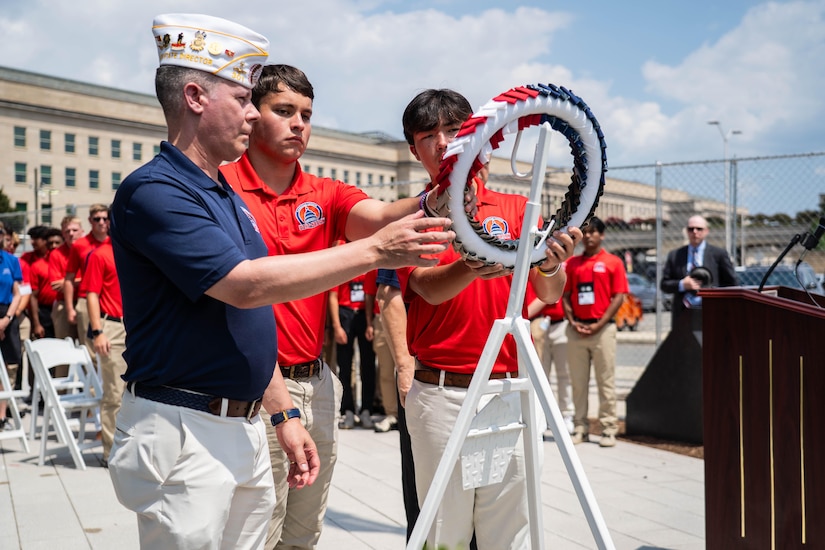 A service member and two students in red polos place a wreath on a white stand outside on a sunny day.