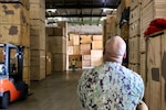 A sailor dressed in a military uniform has his back to the camera as he inspects large boxes in a warehouse.