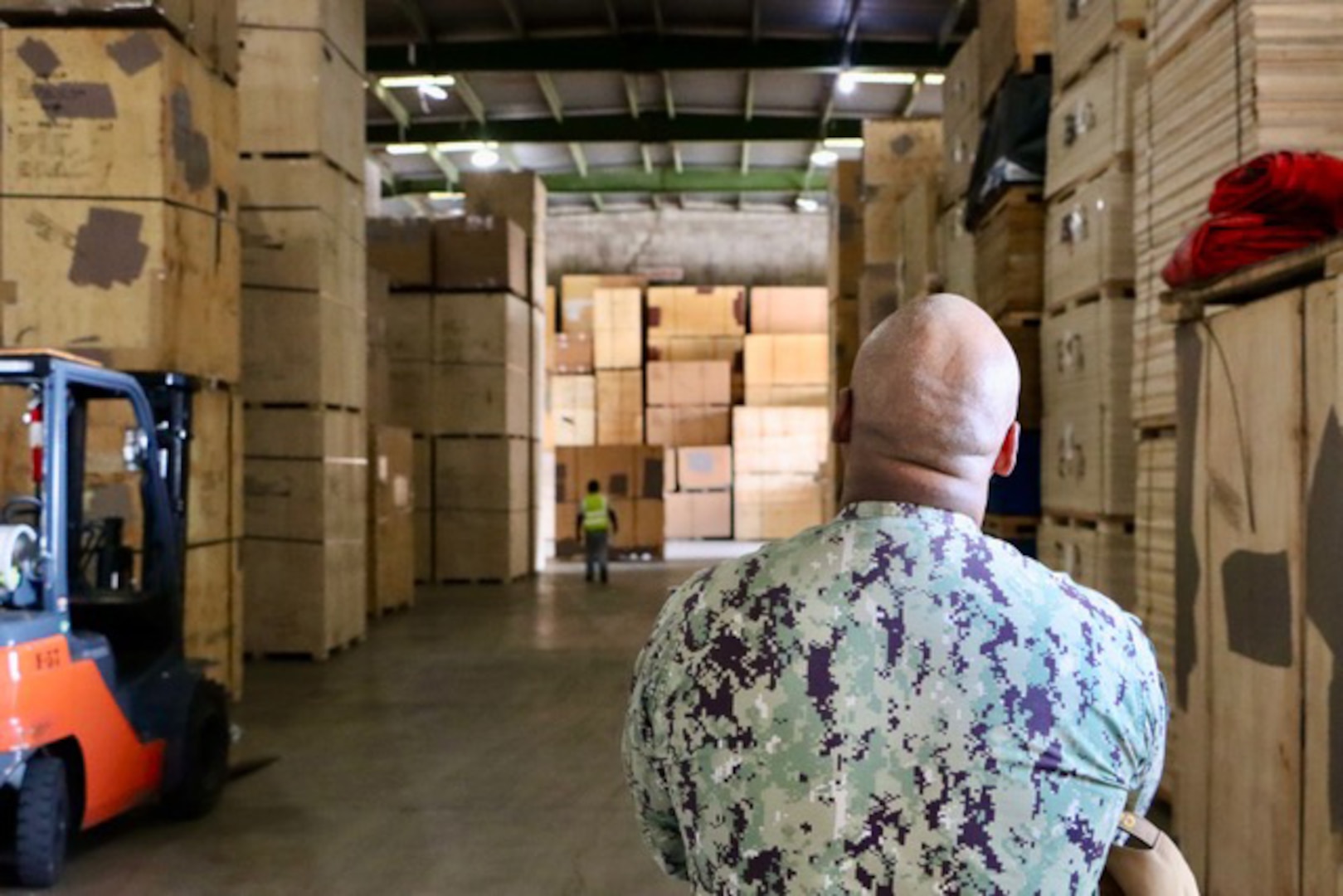 A sailor dressed in a military uniform has his back to the camera as he inspects large boxes in a warehouse.