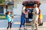 A man and woman unload a suitcase from a car as a child holding a baby watches.