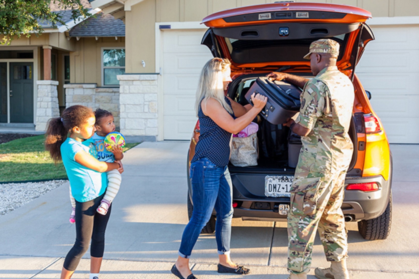 A man and woman unload a suitcase from a car as a child holding a baby watches.