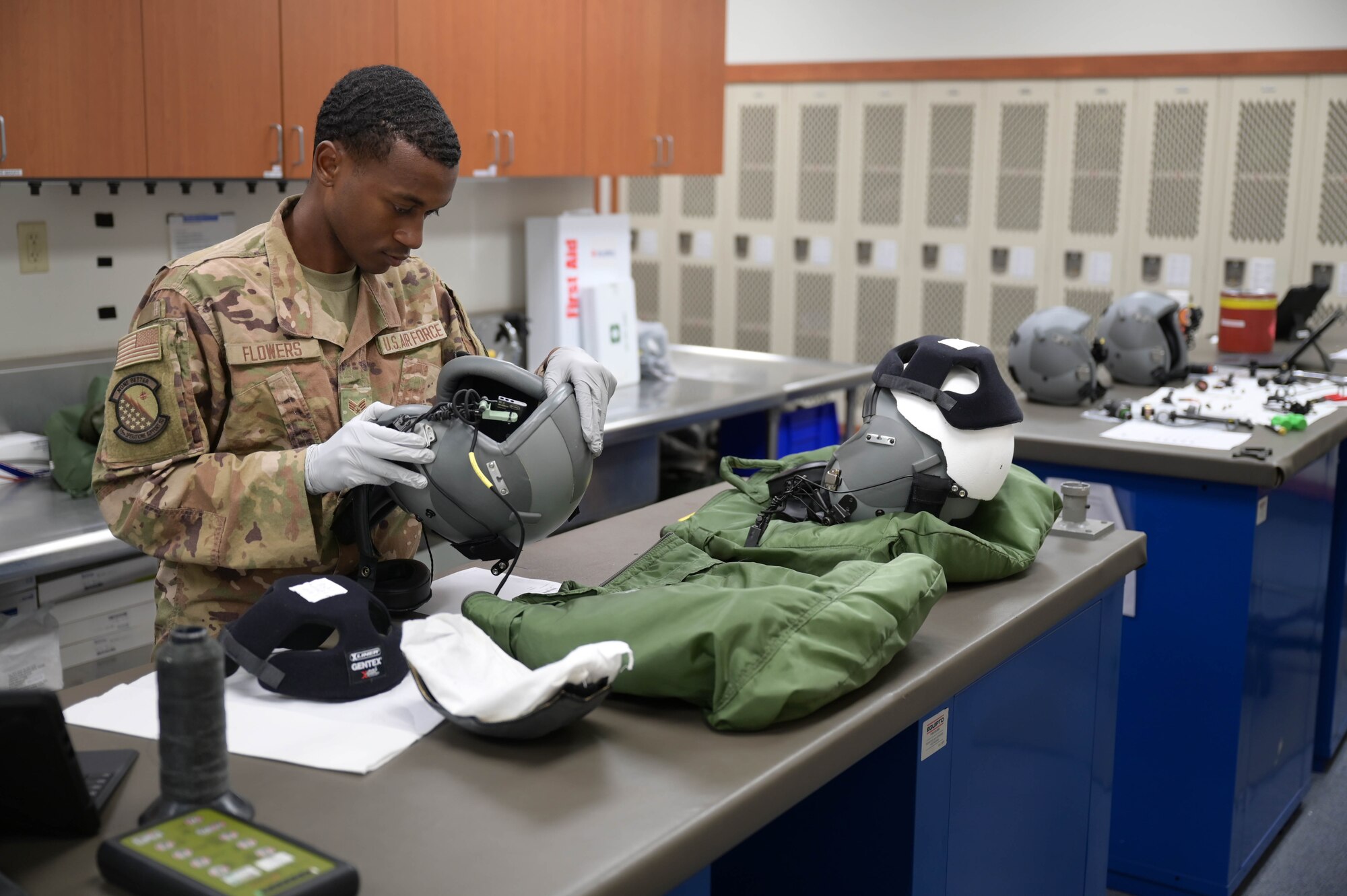 A large room with several pilot helmets laid out on a table with an Airman inspecting one with gloved hands.