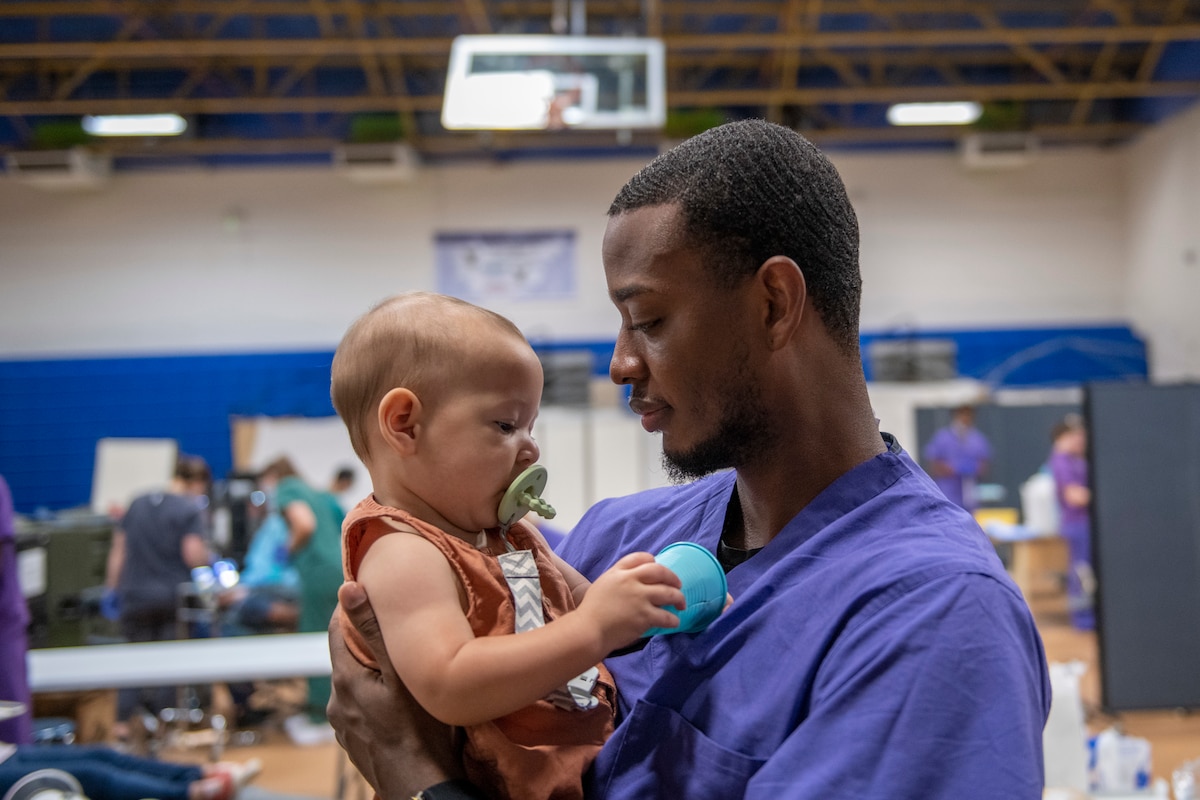 U.S. Air Force Senior Airman Shannando Harrison, a dental technician assigned to the 88th dental squadron, Oh., holds a baby while the baby’s mother receives dental care during Operation Healthy Tennessee, Bledsoe County High School, Pikeville, Tenn., July 18, 2025. Operation Healthy Tennessee provides no-cost medical, dental, vision and veterinary services to the residents of Bledsoe and Rhea County, as well as the surrounding areas while satisfying training requirements for active-duty, reserve and Air National Guard service members and units. (U.S. Air National Guard photo by SSgt. Sarah Stalder Lundgren)