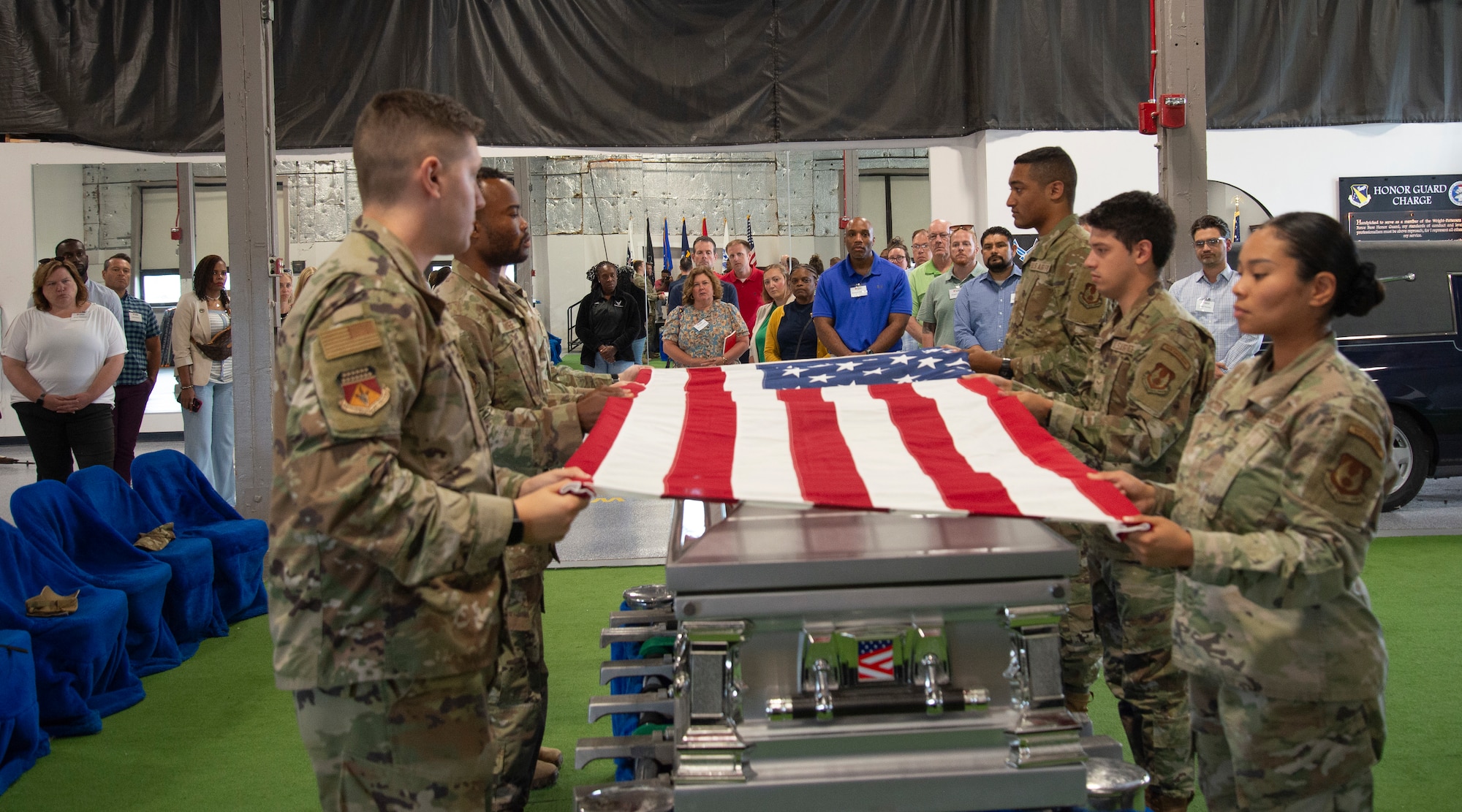 Military members are holding a flag. Preparing to a fold it in front of a group of individuals.