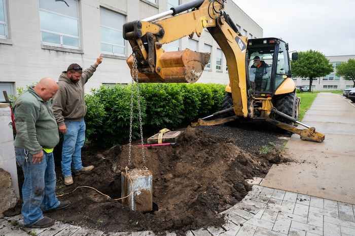 Earth moving equipment pulls heavy time capsule from ground while two workmen guide it out safely.
