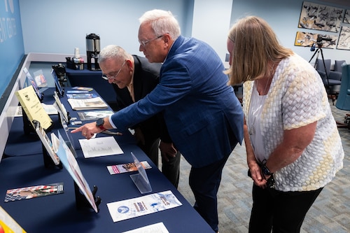 Two men and one woman stand over blue table, looking at brochures.