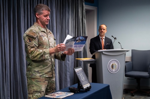 Man in Air Force uniform stands and reads an old newspaper out loud. Man behind podium looks on.