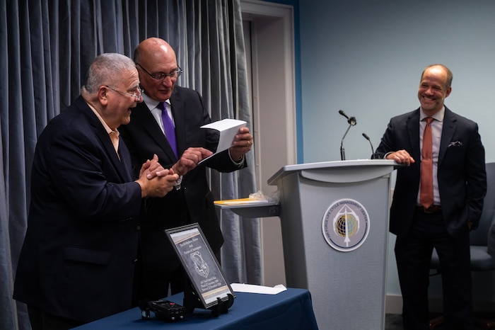 Two men in business suits reading a document together. Man standing beside podium and laughing.