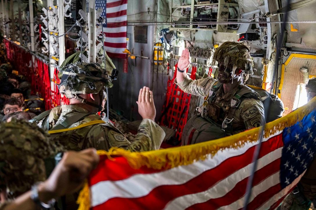 Two soldiers in parachute gear face each other and raise their right hands inside an aircraft while others hold an American flag.