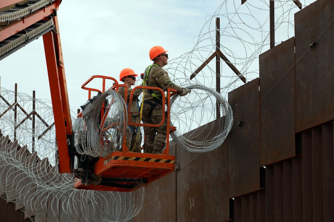 Two soldiers on a red lift install concertina wire along a wall outside.