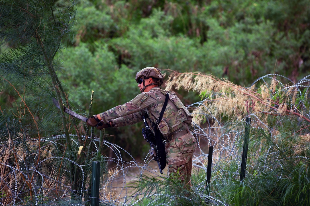 A soldier clears brush from concertina wire outside with a forest behind.