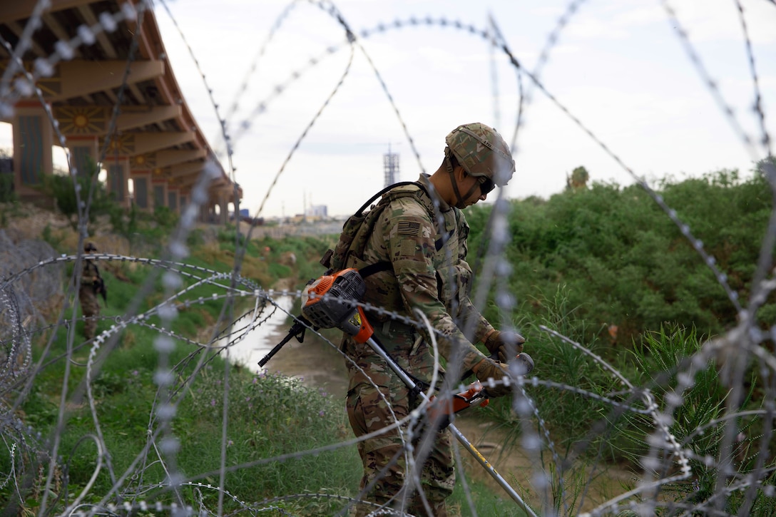 A soldier clears brush next to concertina wire outside on a cloudy day.