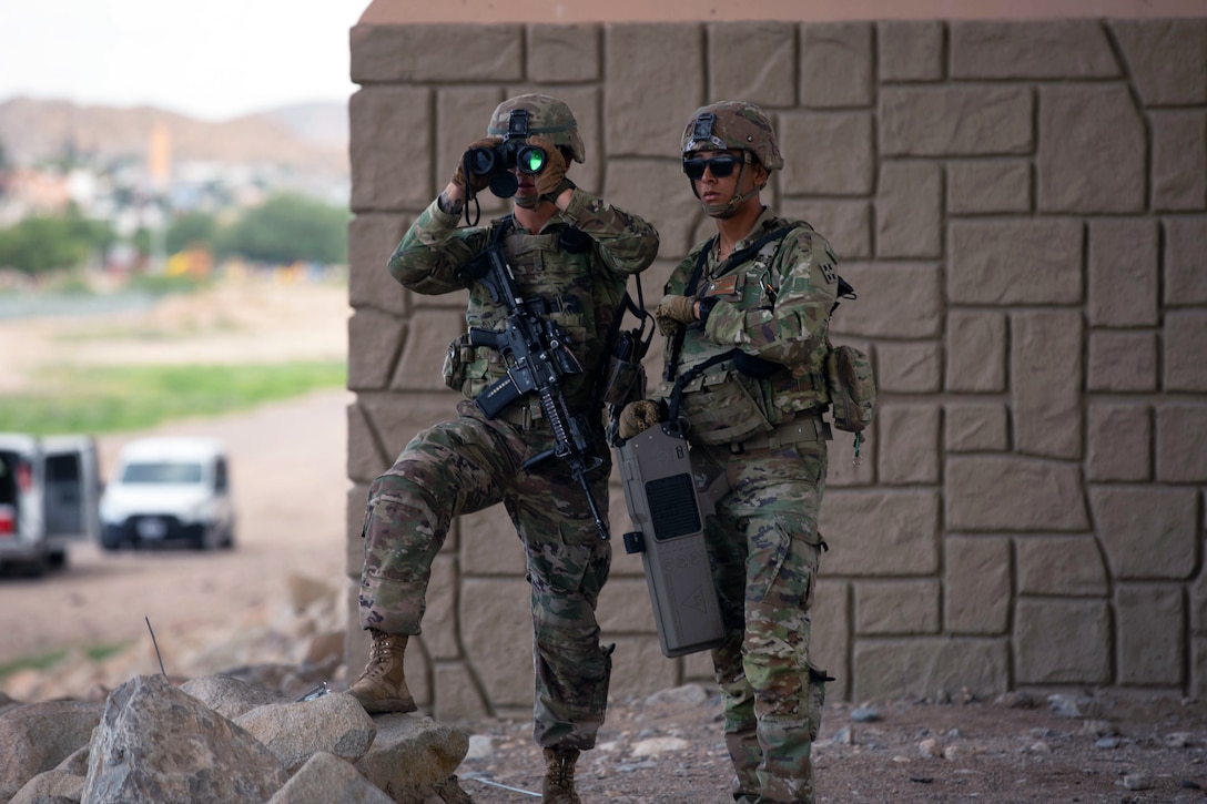 Two soldiers in tactical gear stand by a wall while one uses binoculars.